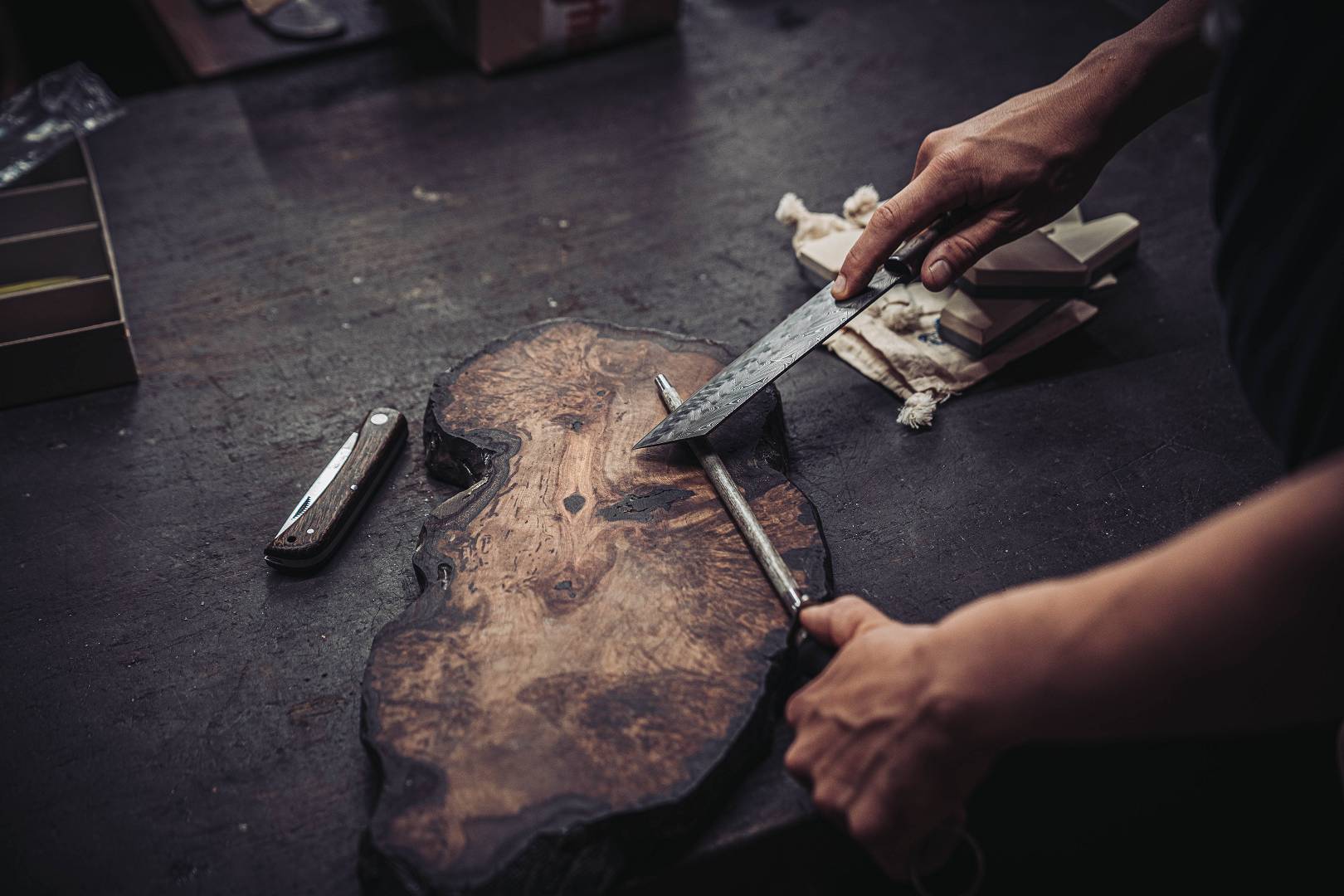 H&auml;nde halten ein Damastmesser &uuml;ber einer Holzscheibe beim Griffanpassen in der Moorschmiede
