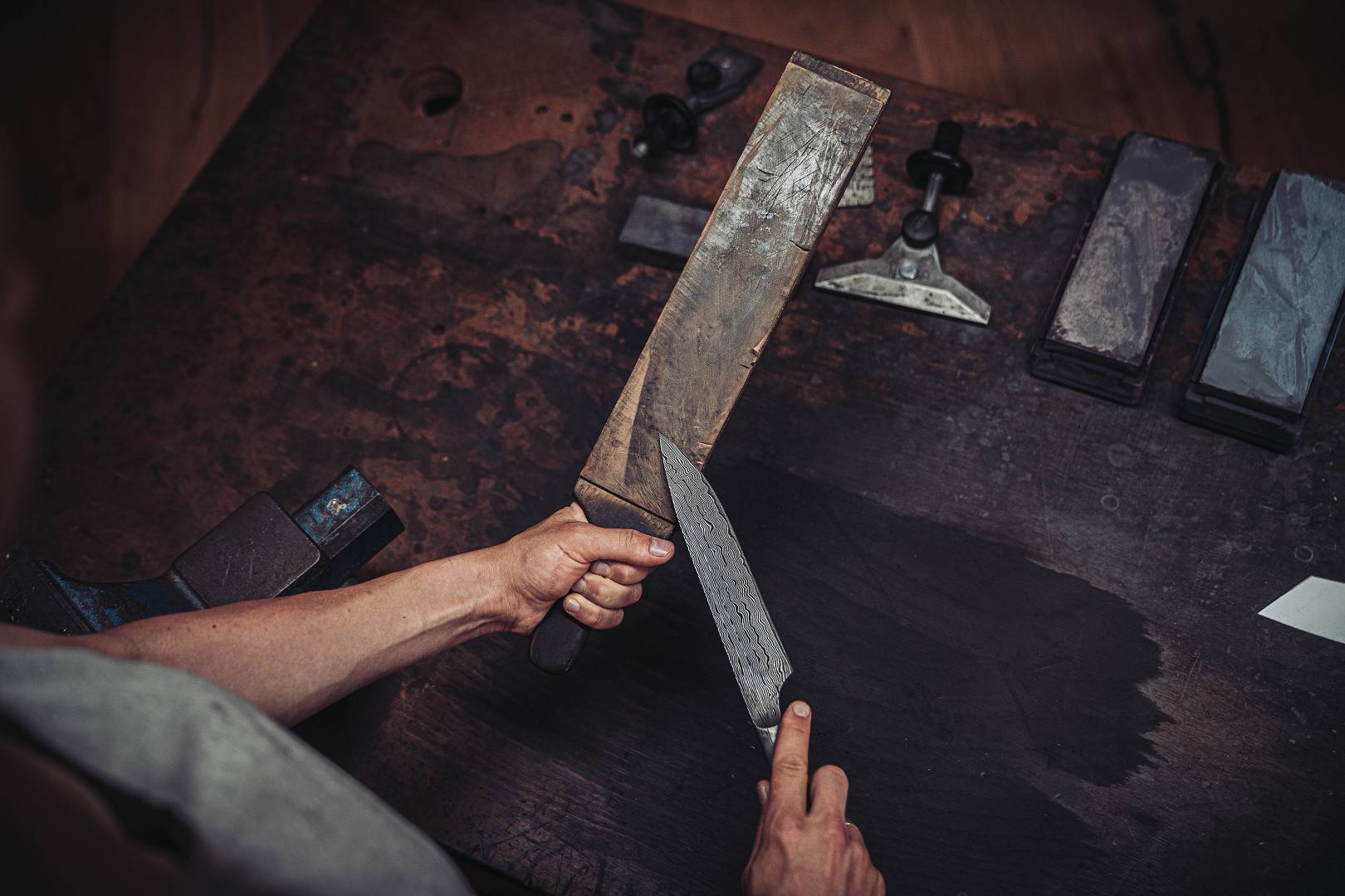 Damastklinge mit sichtbarem Wellenmuster auf Holzblock neben Schleifsteinen